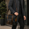 Person holding a brown leather bag wearing a dark coat and pants outdoors.